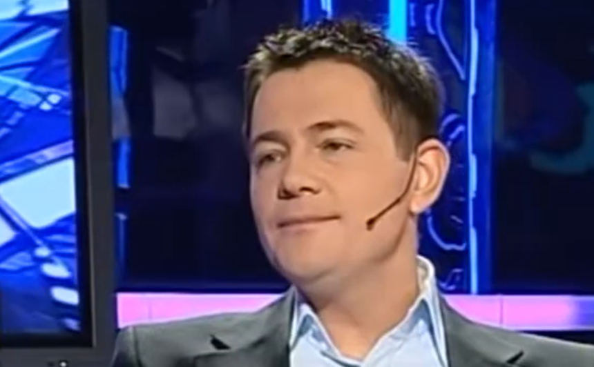 Krzysztof Ibisz z mikrofonem przedstawiony na tle ekranów, uśmiechający się podczas rozmowy.