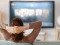 Kobieta siedząca na kanapie, z rękami za głową, ogląda telewizję w przytulnym wnętrzu.