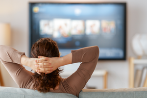Kobieta siedząca na kanapie, z rękami za głową, ogląda telewizję w przytulnym wnętrzu.