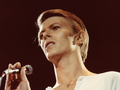 David Bowie. Młody mężczyzna z mikrofonem w ręku, rozświetlony światłem, na tle ciemnej sceny.