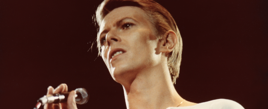 David Bowie. Młody mężczyzna z mikrofonem w ręku, rozświetlony światłem, na tle ciemnej sceny.