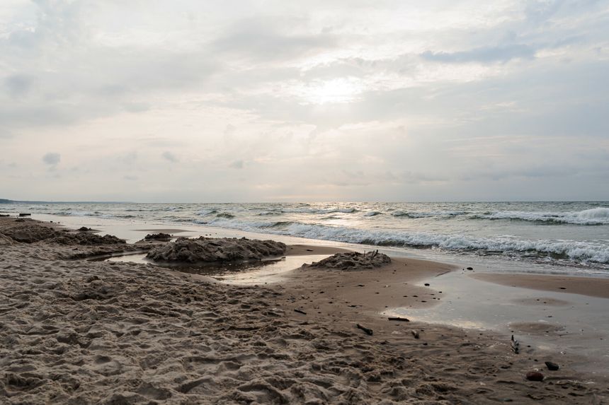 Plaża o zachodzie słońca, fale uderzają w piasek, chmury na niebie. Brak ludzi w kadrze.