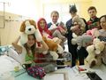 Grupa osób w przebraniach trzyma miśki, odwiedzając dzieci w szpitalu. W centrum dziewczynka z zabawką.