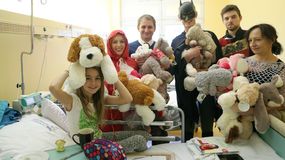 Grupa osób w przebraniach trzyma miśki, odwiedzając dzieci w szpitalu. W centrum dziewczynka z zabawką.