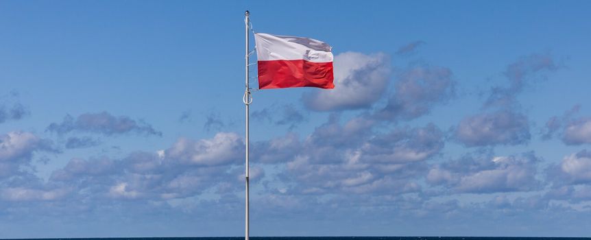 Flaga Polski na tle nieba i morza, w otoczeniu białych chmur.