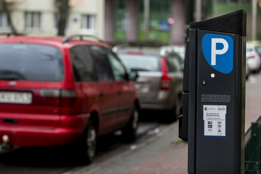 Automat parkingowy na ulicy z zaparkowanymi samochodami w tle. 