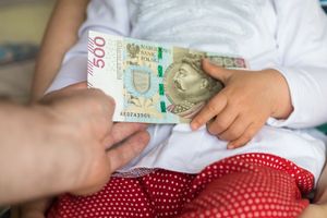 Dziecko trzyma banknot 500 złotych, drugą ręką przytrzymując rękę dorosłego.