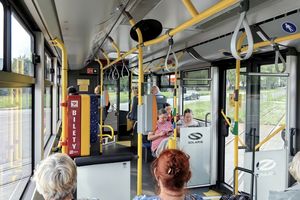 Wnętrze autobusu miejskiego z pasażerami siedzącymi i stojącymi, widoczny biletomat.