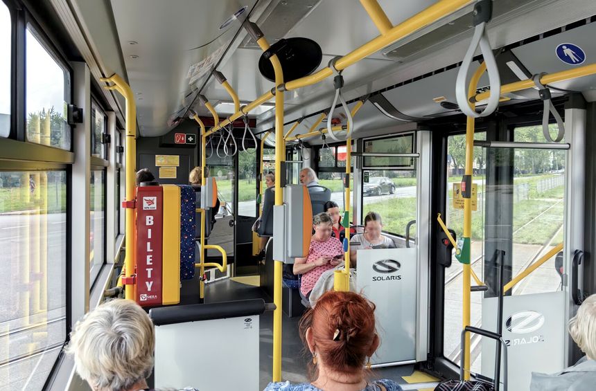 Wnętrze autobusu miejskiego z pasażerami siedzącymi i stojącymi, widoczny biletomat.
