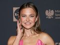 Anna Lewandowska w różowej sukience uśmiecha się, dotykając dłonią twarzy.