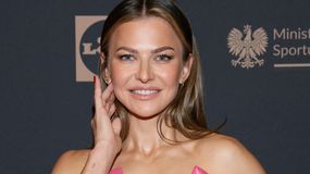 Anna Lewandowska w różowej sukience uśmiecha się, dotykając dłonią twarzy.