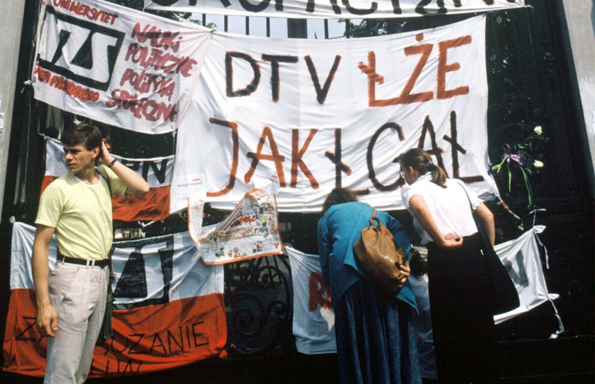 Grupa ludzi przed budynkiem, z transparentami i flagami, protestujących w ważnej sprawie.