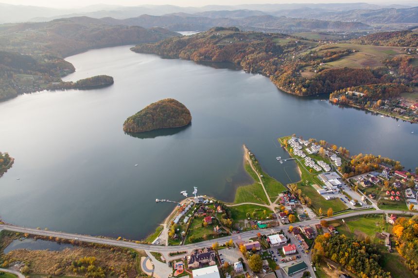 Widok na jezioro z wyspą, otoczone wzgórzami i domami w jesiennych kolorach.