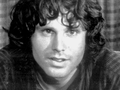 Jim Morrison. Mężczyzna z długimi, kręconymi włosami i uśmiechem, siedzący na tle kratkowanej zasłony.