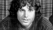 Jim Morrison. Mężczyzna z długimi, kręconymi włosami i uśmiechem, siedzący na tle kratkowanej zasłony.