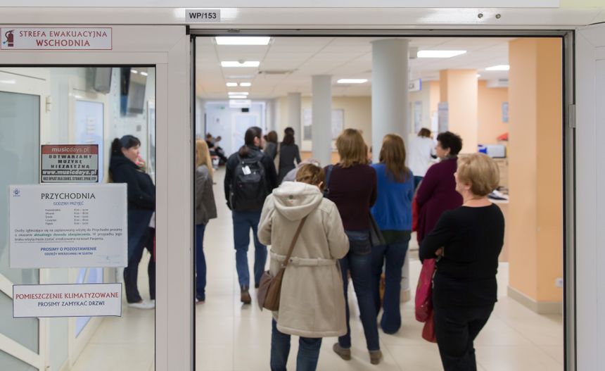 Opis ALT: Długi korytarz przychodni, kolejka pacjentów czekających na wizytę.