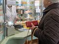Osoba starsza w sklepie aptecznym rozmawia z farmaceutą za szybą. Trzyma czerwoną torebkę.