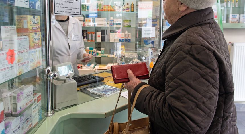Osoba starsza w sklepie aptecznym rozmawia z farmaceutą za szybą. Trzyma czerwoną torebkę.