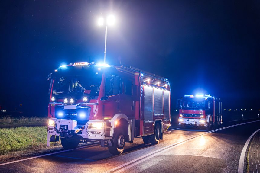 Na zdjęciu widoczne są dwa czerwone wozy strażackie z niebieskimi i białymi światłami na drodze nocą.