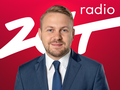 Mężczyzna w garniturze na tle czerwonym z napisem „radio ZłT”. Uśmiecha się do kamery.