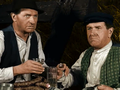 Dwaj mężczyźni w kapeluszach siedzą przy stole, trzymając szklanki z alkoholem. W tle siano.