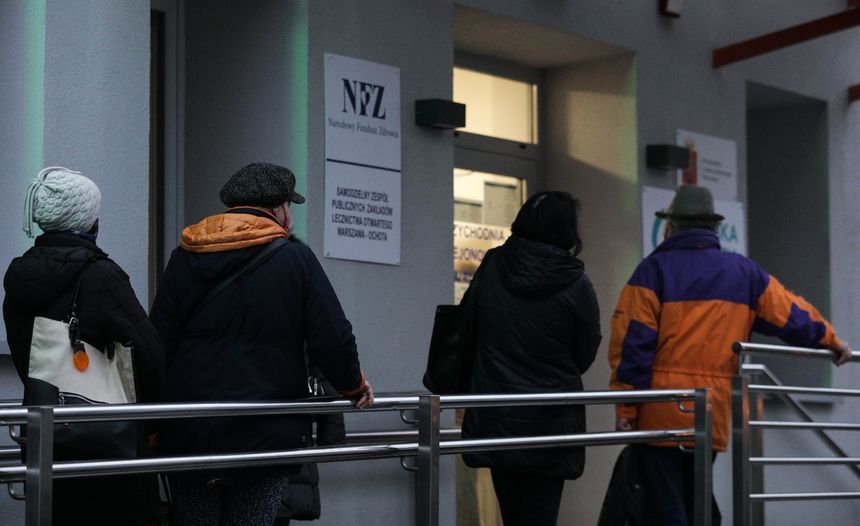 Na zdjęciu cztery osoby stojące w kolejce do budynku NFZ w chłodny dzień.