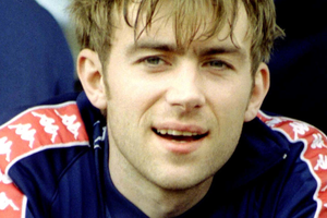 Damon Albarn z zespołu Blur. Mężczyzna z krótkimi włosami, uśmiechający się w sportowym stroju, siedzi na ławce.