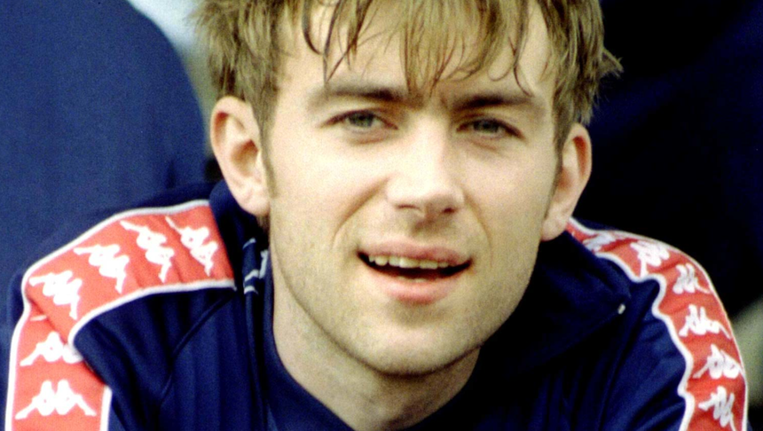 Damon Albarn z zespołu Blur. Mężczyzna z krótkimi włosami, uśmiechający się w sportowym stroju, siedzi na ławce.