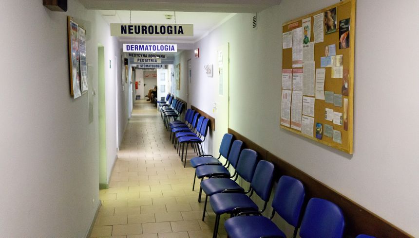 Korytarz przychodni z niebieskimi krzesłami i drogowskazami do lekarzy specjalistów.