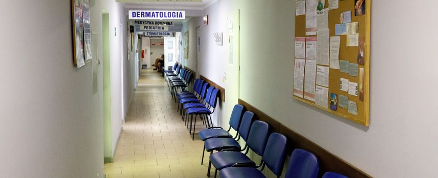 Korytarz przychodni z niebieskimi krzesłami i drogowskazami do lekarzy specjalistów.
