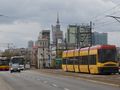 Zdjęcie przedstawia tramwaj i autobusy na ruchliwej ulicy, w tle wieżowiec i chmury.