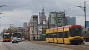 Zdjęcie przedstawia tramwaj i autobusy na ruchliwej ulicy, w tle wieżowiec i chmury.
