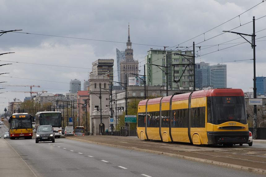 Zdjęcie przedstawia tramwaj i autobusy na ruchliwej ulicy, w tle wieżowiec i chmury.