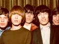 The Rolling Stones. Pięć osób w stylowych ubraniach, pozujących na tle zasłony. Radość i uśmiechy.