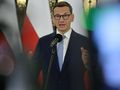 Mężczyzna w okularach, mówiący do mikrofonu, z flagami w tle. 