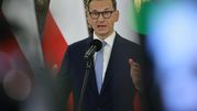 Mężczyzna w okularach, mówiący do mikrofonu, z flagami w tle. 