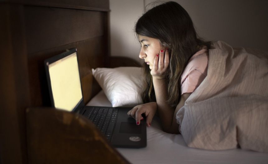 Dziewczynka leży na łóżku, skupiona na laptopie w ciemnym pokoju, z ręką na twarzy.