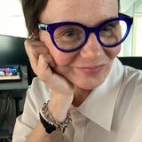 Kobieta z krótkimi włosami, niebieskimi okularami, uśmiecha się, siedząc przy biurku.