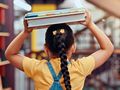 Dziewczynka w żółtej bluzce i z warkoczem niesie stos książek w bibliotece.
