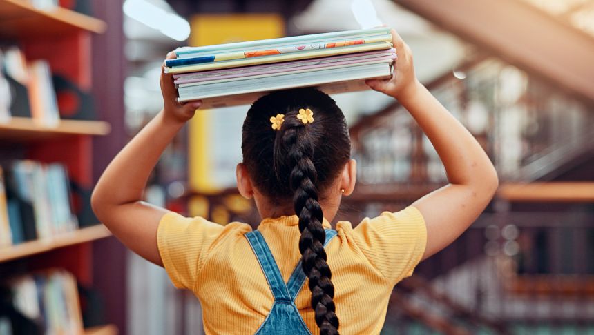 Dziewczynka w żółtej bluzce i z warkoczem niesie stos książek w bibliotece.
