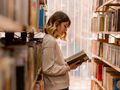 Młoda kobieta stoi między regałami bibliotecznymi, trzymając książkę i czytając ją z zainteresowaniem.