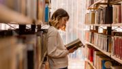 Młoda kobieta stoi między regałami bibliotecznymi, trzymając książkę i czytając ją z zainteresowaniem.