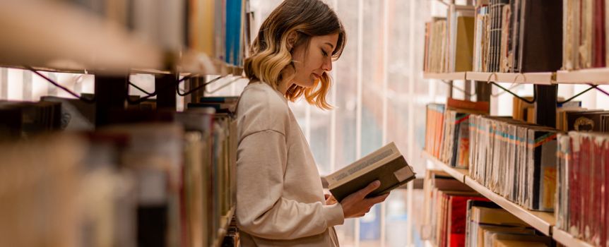 Młoda kobieta stoi między regałami bibliotecznymi, trzymając książkę i czytając ją z zainteresowaniem.
