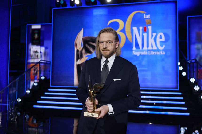 Mężczyzna w garniturze trzyma statuetkę Nagrody Nike na tle niebieskiej sceny.