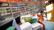 Wnętrze apteki z półkami pełnymi leków, widoczna farmaceutka obsługująca klientów.