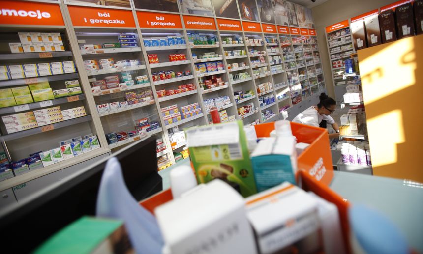 Wnętrze apteki z półkami pełnymi leków, widoczna farmaceutka obsługująca klientów.
