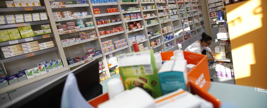 Wnętrze apteki z półkami pełnymi leków, widoczna farmaceutka obsługująca klientów.