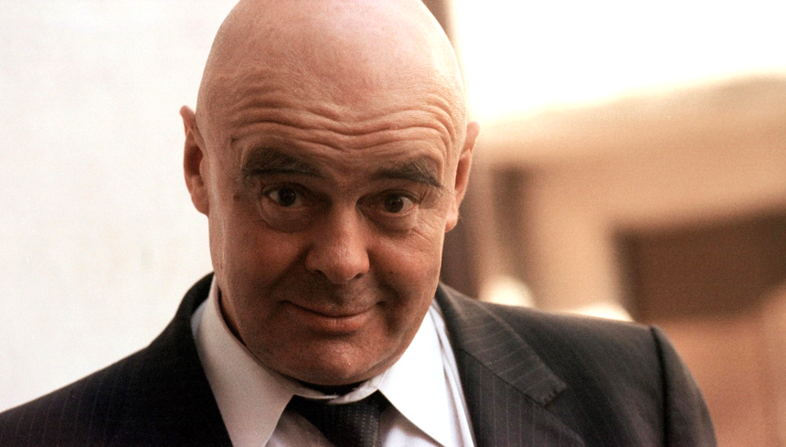 Mężczyzna w garniturze, z łysą głową, uśmiecha się i patrzy w stronę kamery.