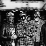 Grupa pięciu mężczyzn w maskach, ubranych w różne stylowe kurtki, na miejskiej ulicy.