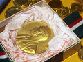 Złoty medal Nobla w ozdobnym pudełku, otoczony kolorowymi monetami.